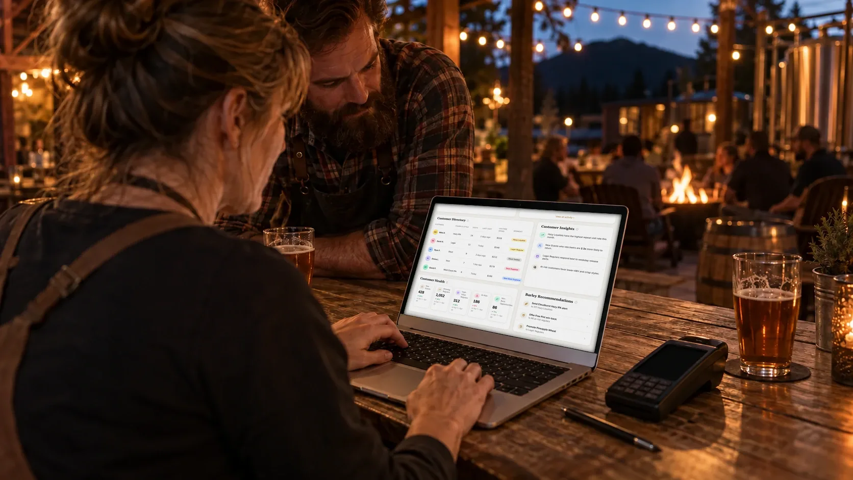 A taproom manager types on a laptop showing the Brewlytics customer directory and customer insights, on a string-lit patio beside a card reader and a pint.