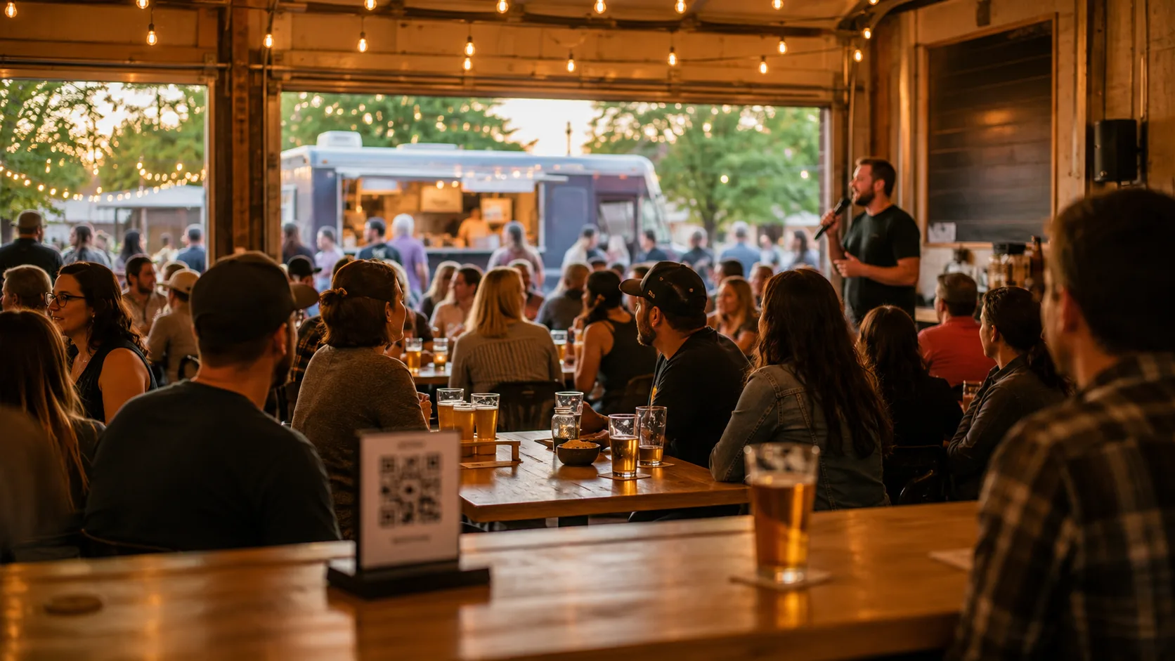 A lively brewery taproom event: customers gathered around tables for trivia night, a host with a microphone, string lights overhead, pint glasses raised, a food truck visible through the open garage door, and a QR code sign on the bar inviting check-ins.