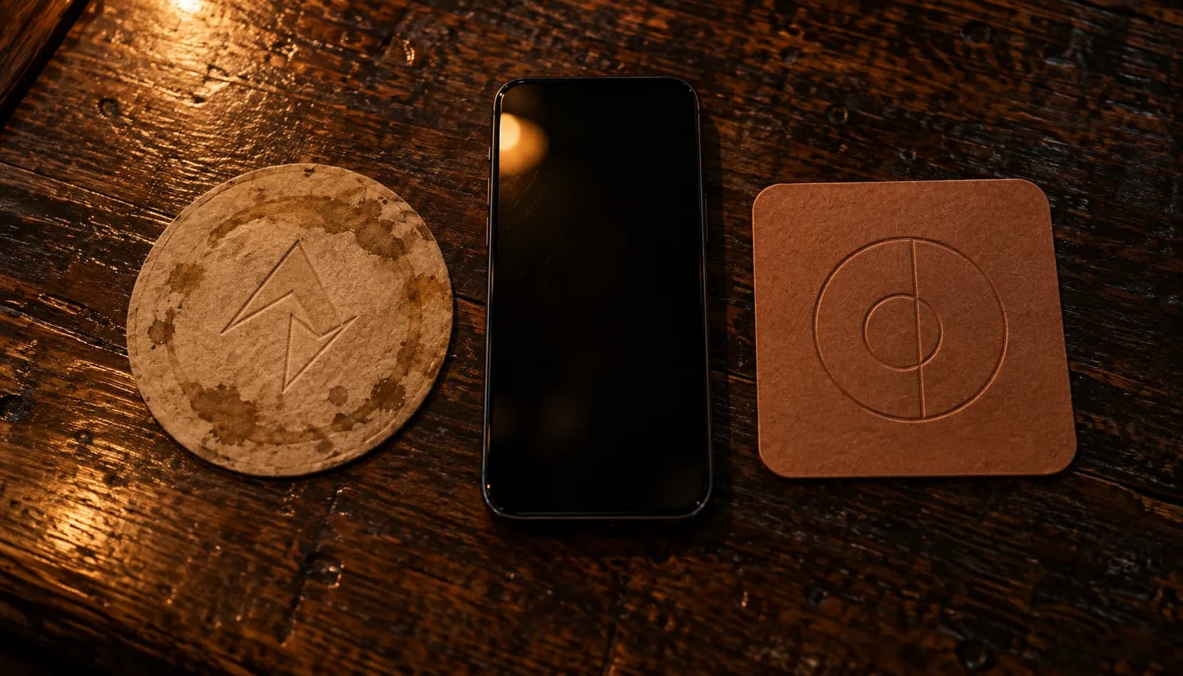 Overhead close-up on a dark wood bar top: two coasters from two different taprooms, one round and ring-stained, the other square and new, lying either side of a single phone with its screen switched off.