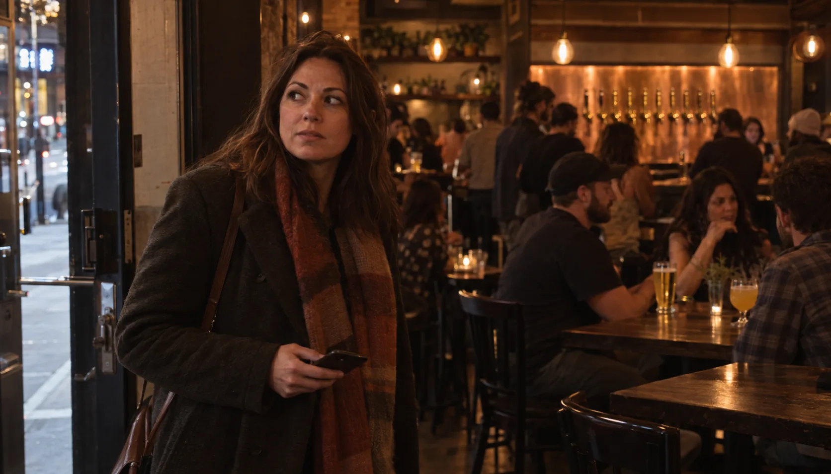 A woman in her thirties with her coat still on, standing just inside the door of a busy downtown taproom on a Thursday night, holding her phone and looking around a room she does not know yet.