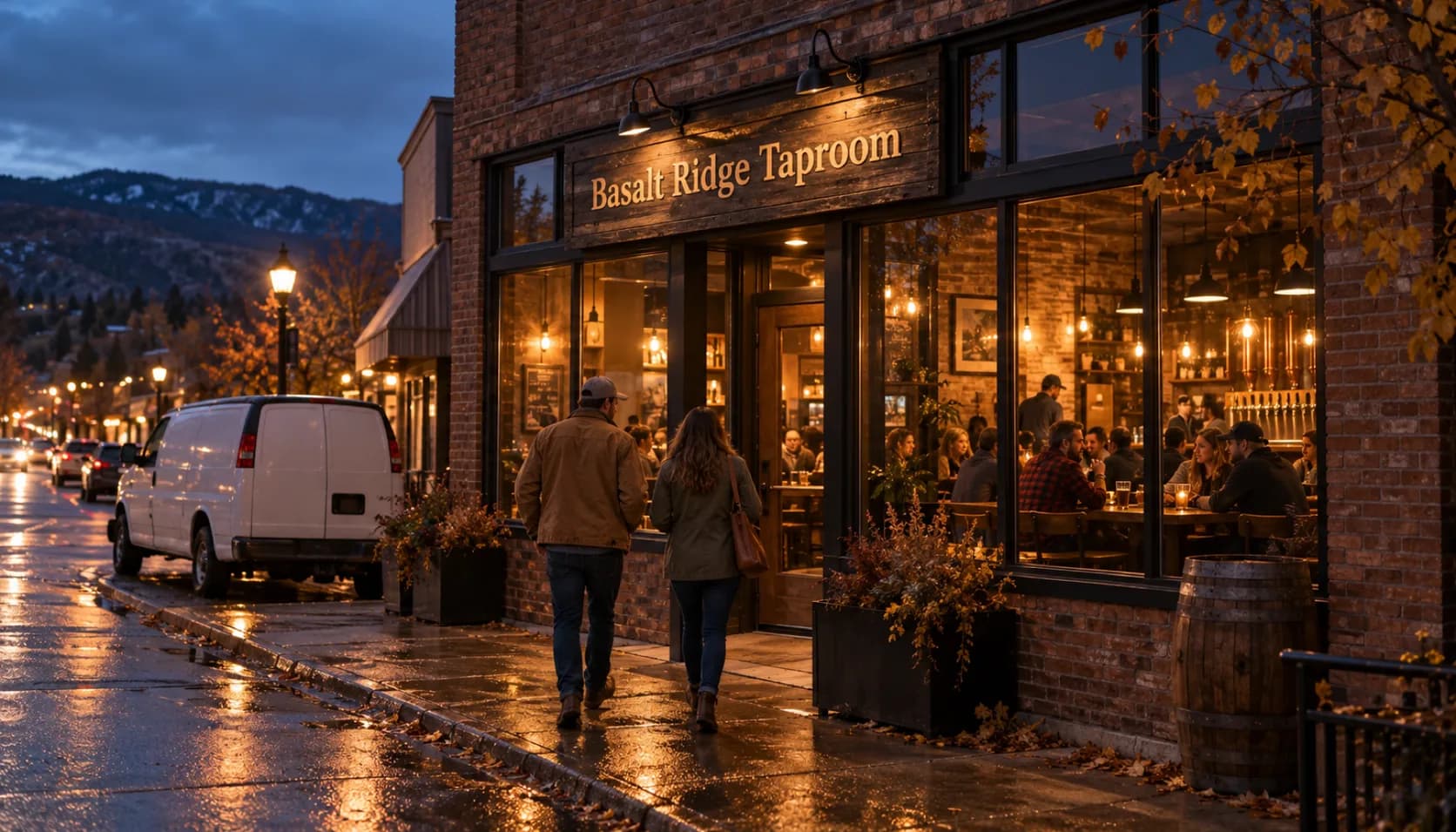 Two people walk along a rain-wet main street at dusk toward a brick storefront taproom with a sign reading Basalt Ridge Taproom, its tall windows glowing over full tables, a plain white van at the curb and hills behind.