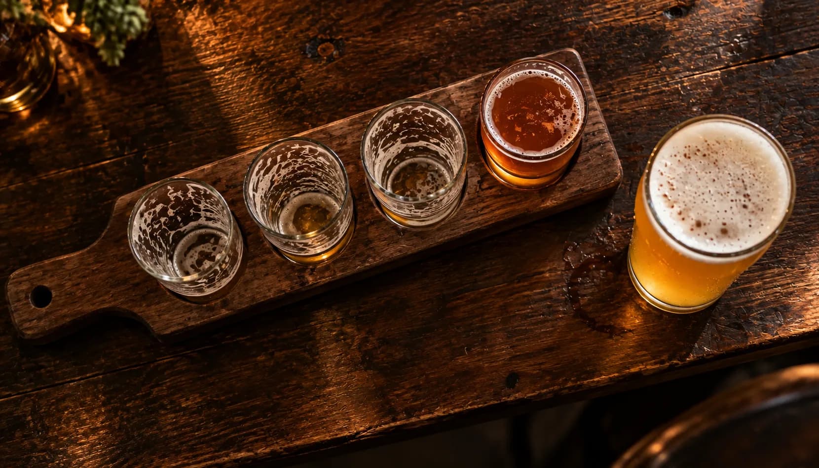 Overhead view of a wooden flight paddle on a dark wood table, three taster glasses drained and one still full of amber beer, beside a full pint of golden beer.