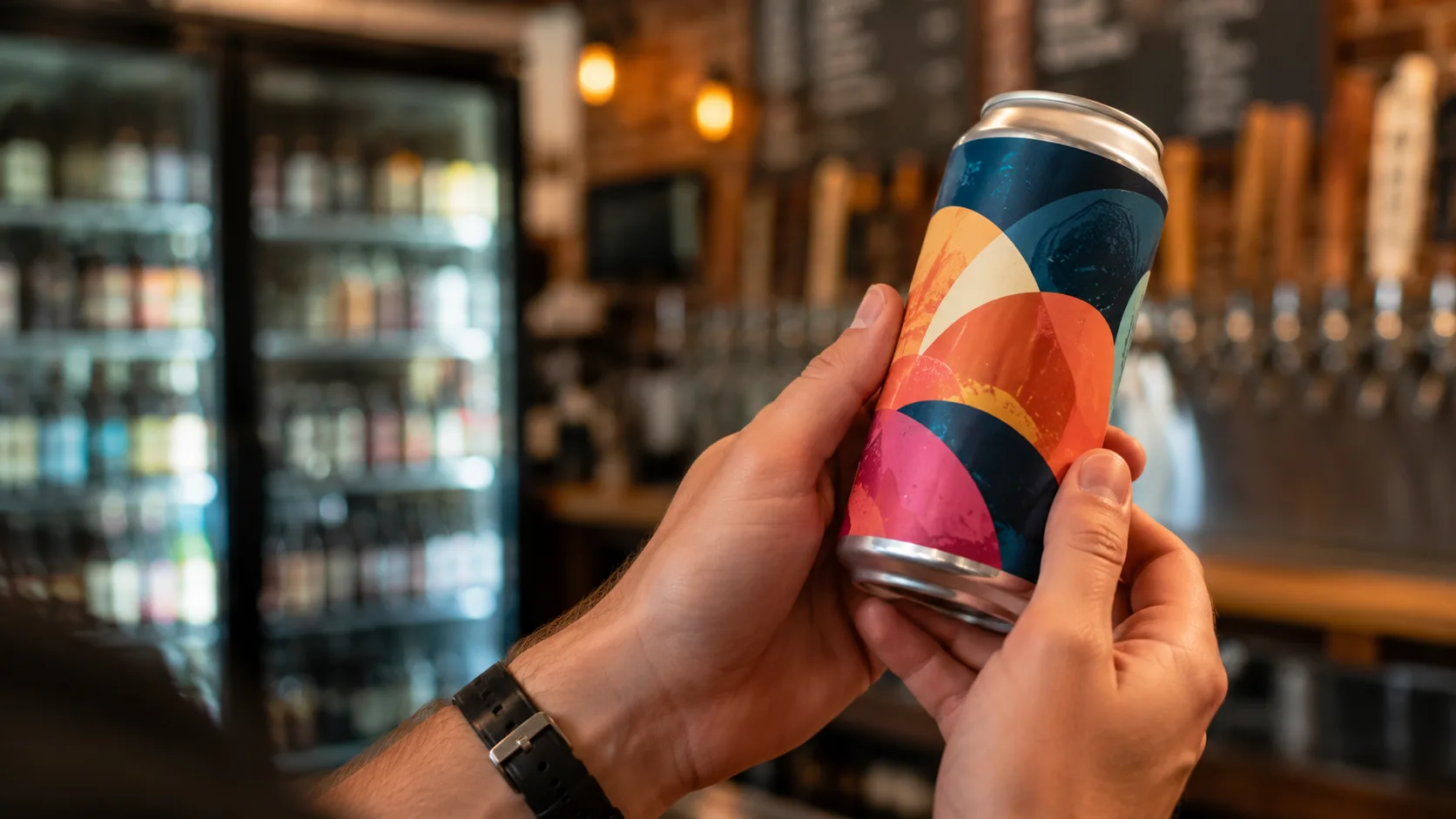 Two hands hold a colorful, unlabeled craft beer can up to inspect it in a warm taproom, with a cooler of bottles blurred in the background.