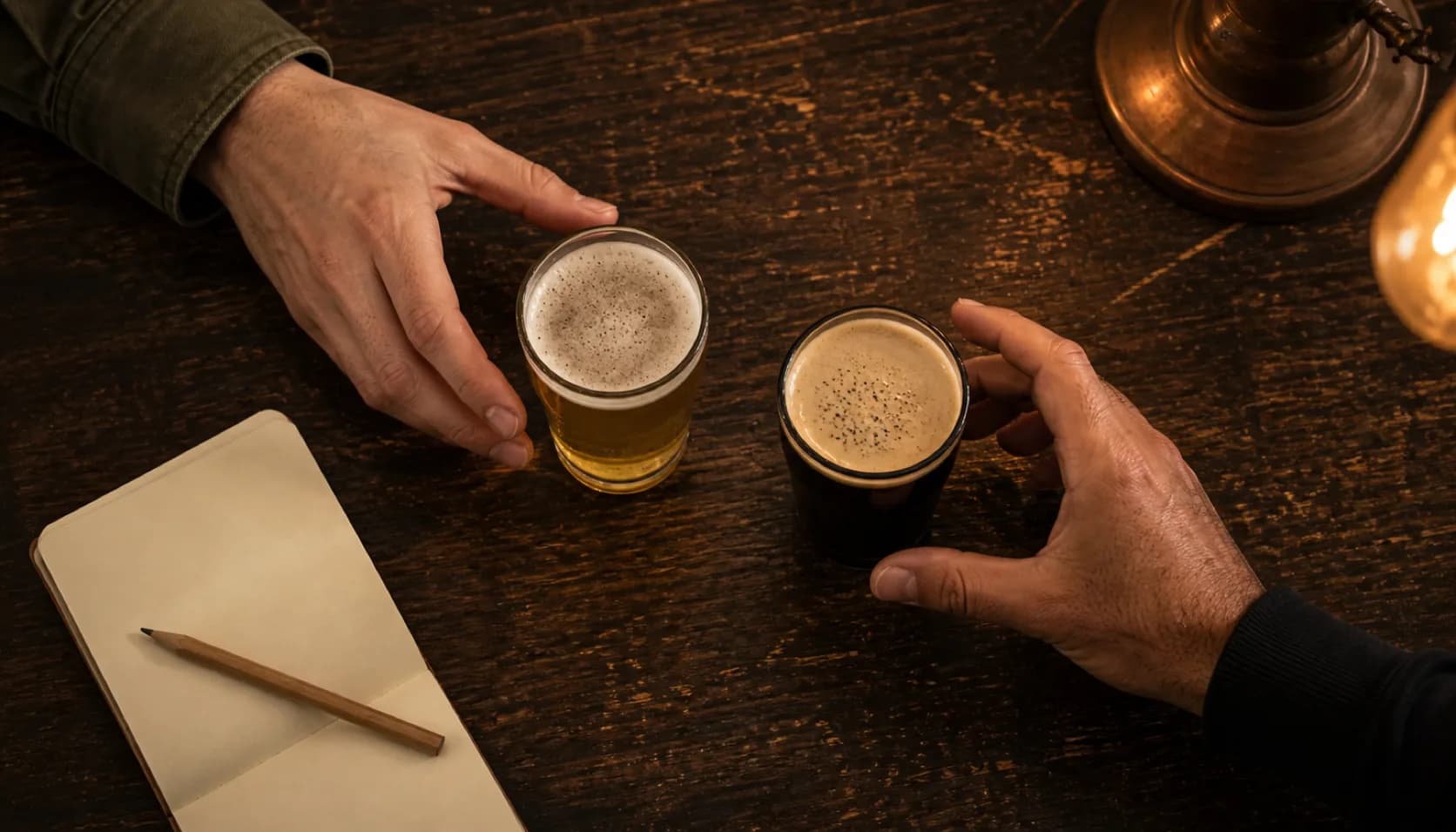 Overhead view of two small tasting pours on a dark wooden table, one pale and one dark, with two people's hands reaching in from opposite sides beside an open notebook and pencil.