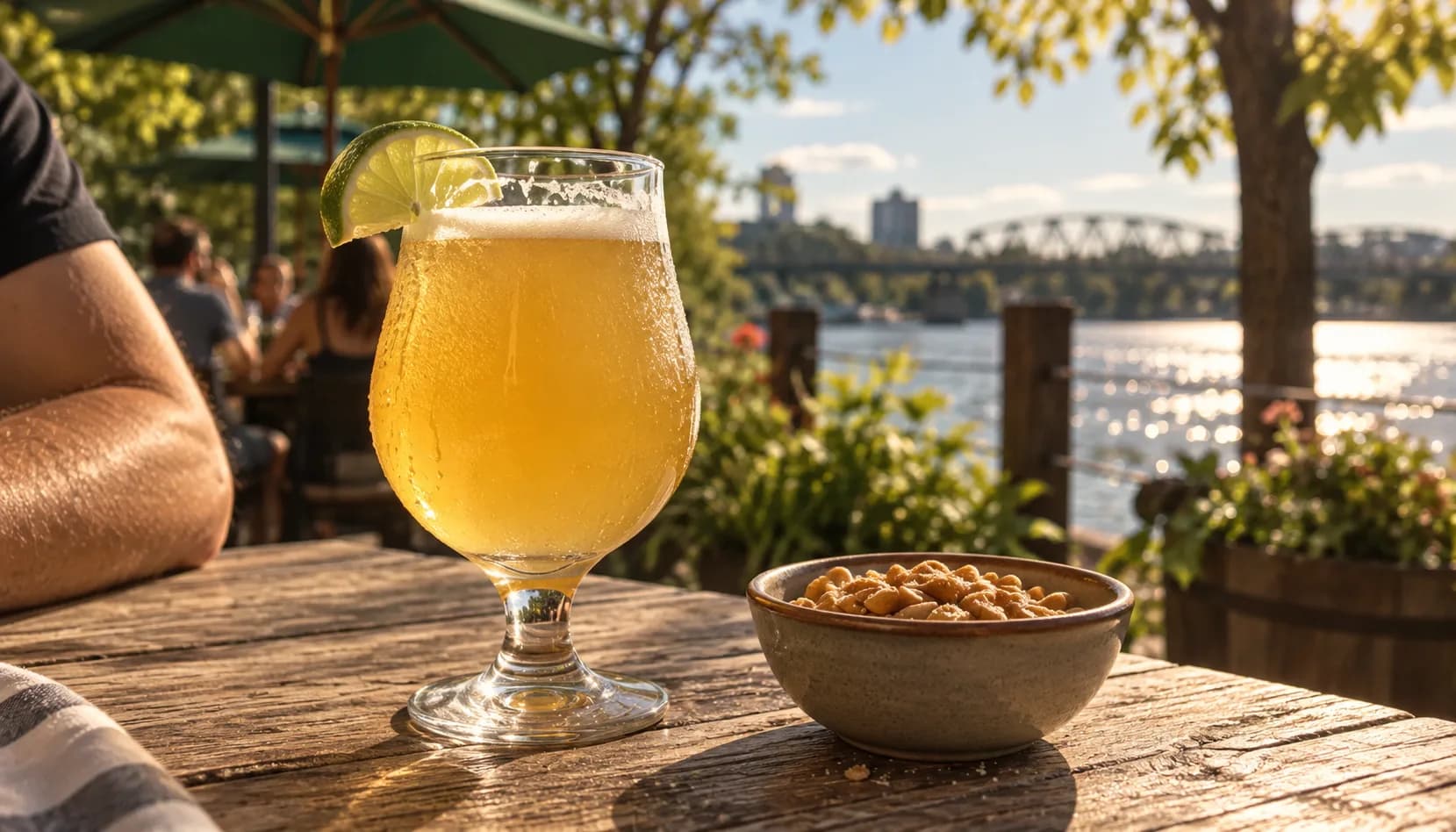 A sweating stemmed glass of pale, hazy gose with a lime wedge on the rim next to a bowl of peanuts on a sunny riverside patio table, with a bridge and bright water in the background.