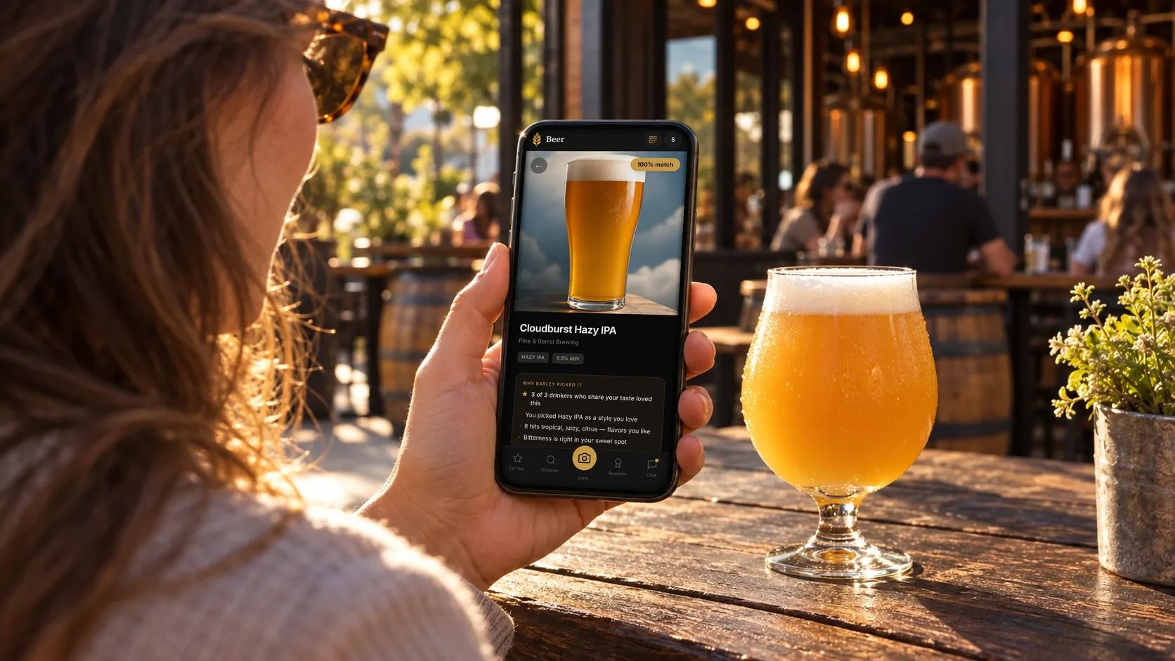 On a sunny patio, a drinker checks Cloudburst Hazy IPA in the free Barley app beside a glowing hazy IPA: a 100% match, and why Barley picked it for her.