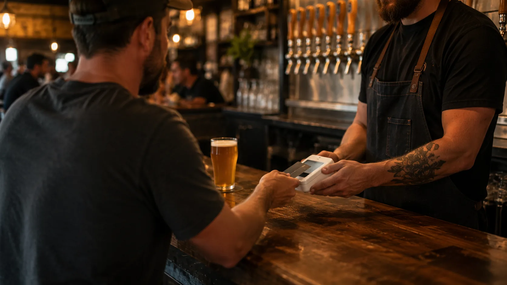 A bartender taking a card payment on a handheld reader at the taproom bar, a fresh pint on the counter.